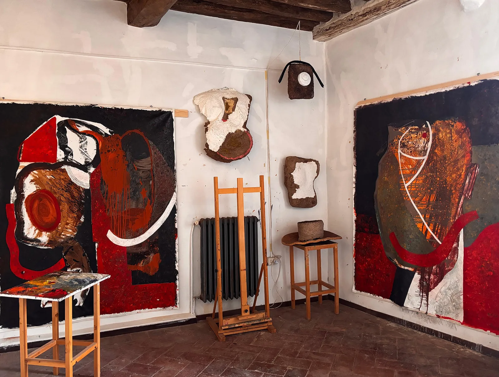 Daria Most's studio: two large abstract paintings in red, black and ochre hang on white walls beside papier-mâché wall pieces, under a wooden beamed ceiling.